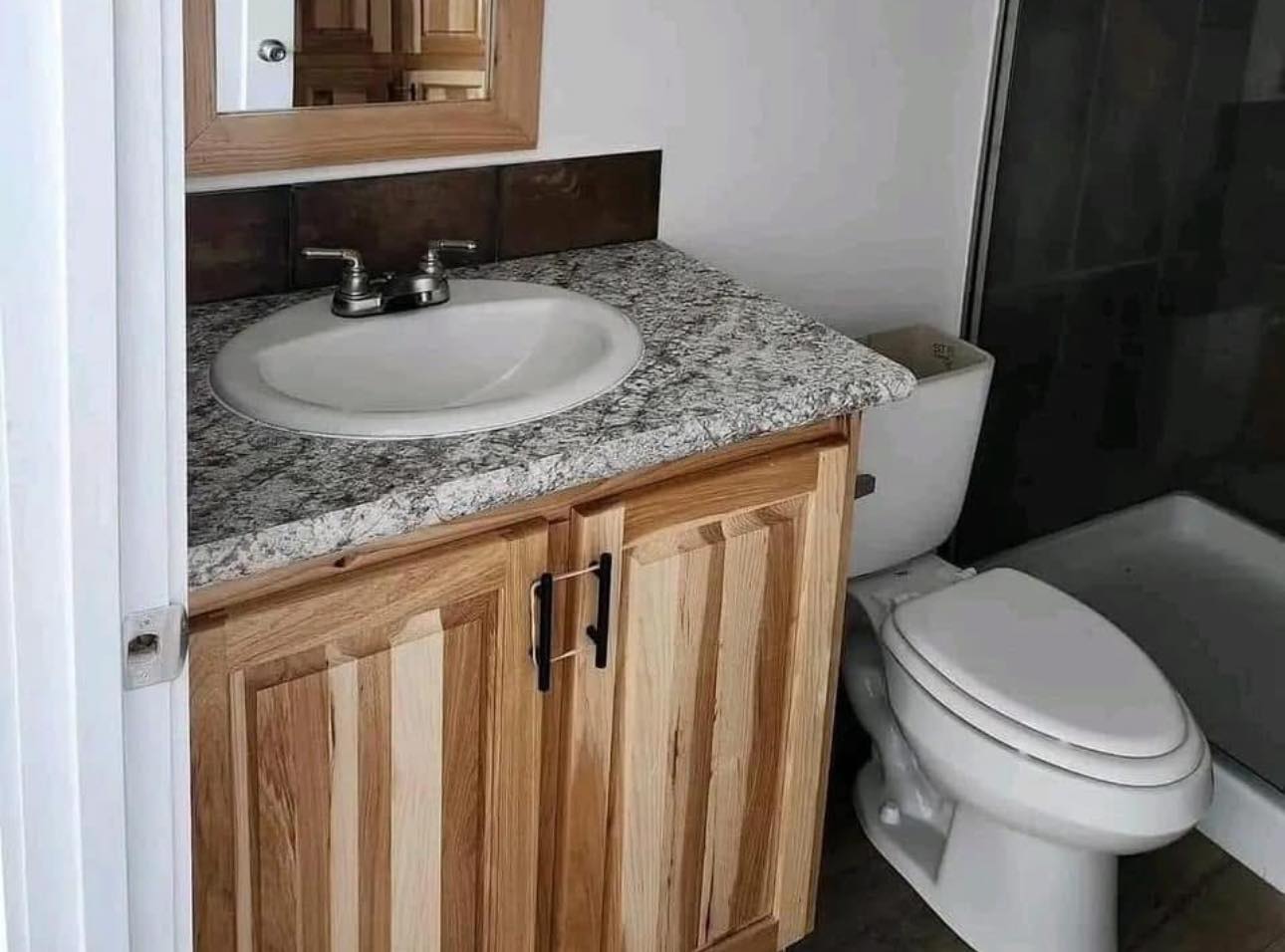 Bathroom with a rustic vibe featuring a granite countertop, wooden vanity, and white sink. Adjacent are a white toilet and a shower stall with dark tiles.