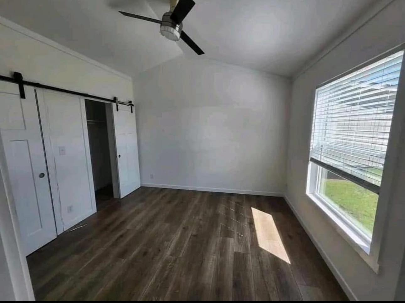 Empty room with light wood flooring, a ceiling fan, and white walls. A large window on the right lets in sunlight, and there's a closet with sliding doors on the left.