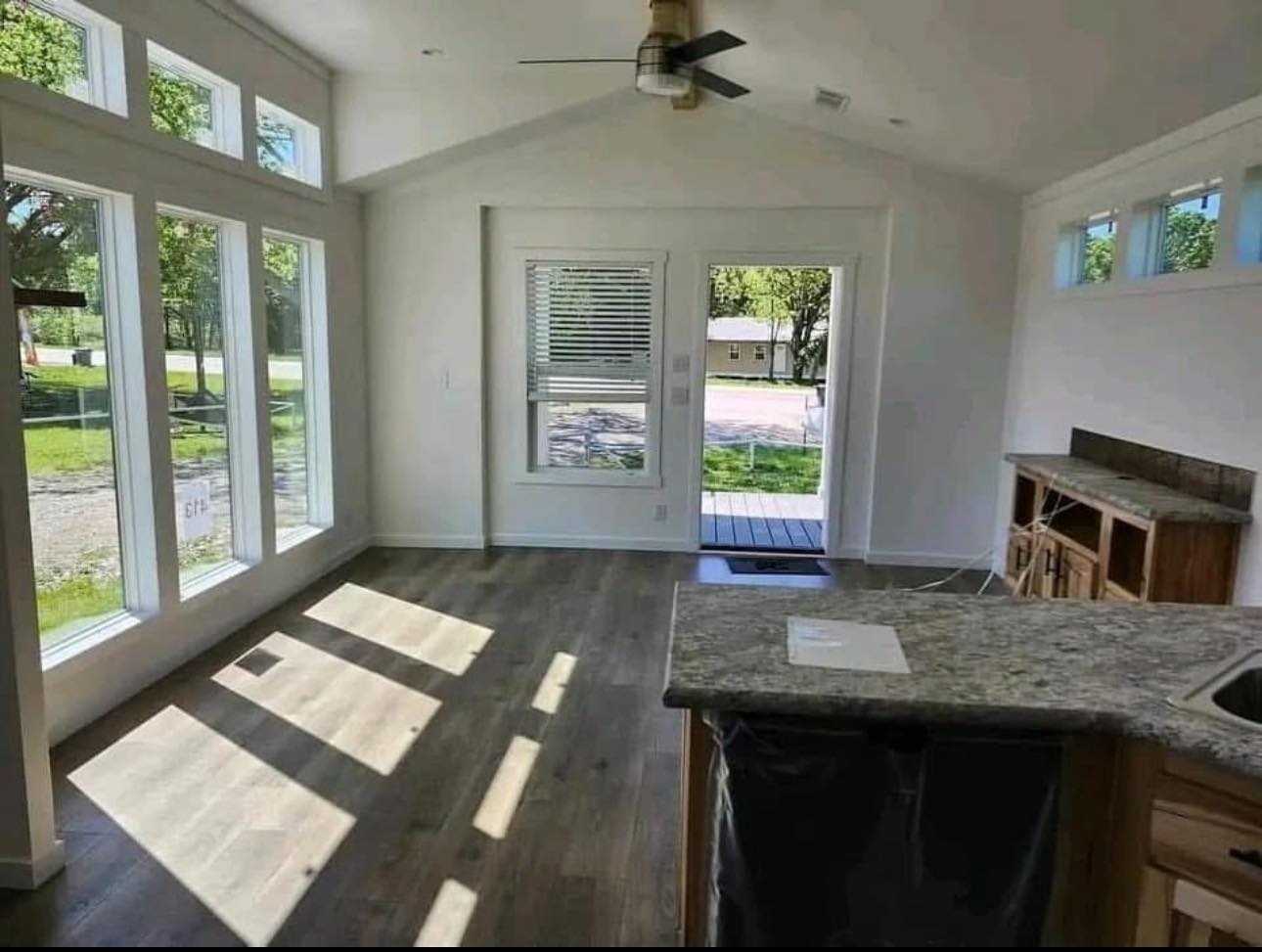 Bright, modern room with large windows, light wood floors, and a small kitchen area. Natural light casts shadows, creating a warm, inviting atmosphere.