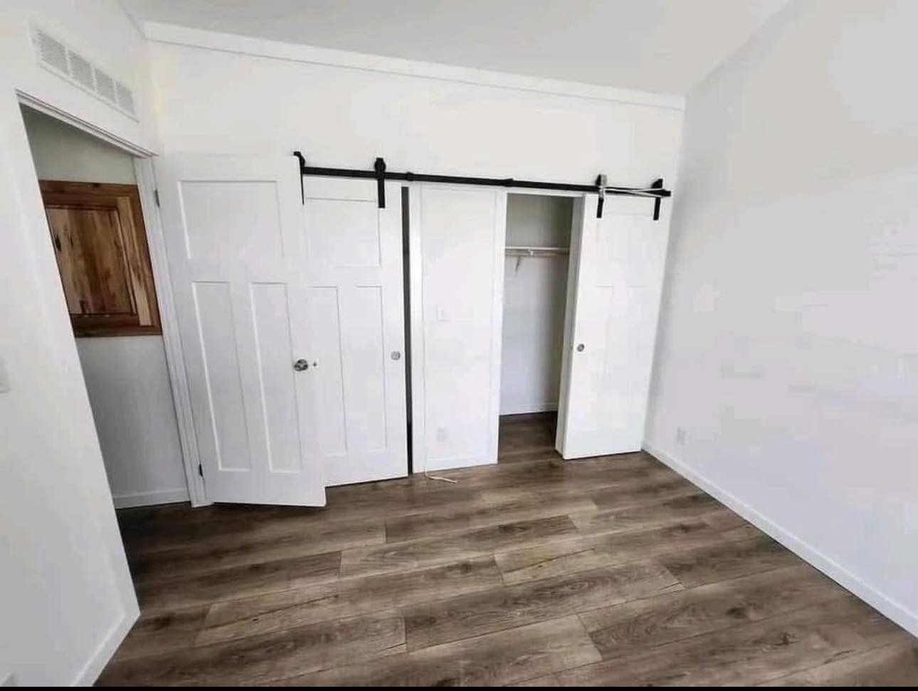 Empty room with wooden flooring, white walls, and a sliding barn door closet. Open closet reveals a hanging rod, creating a minimalist, airy feel.