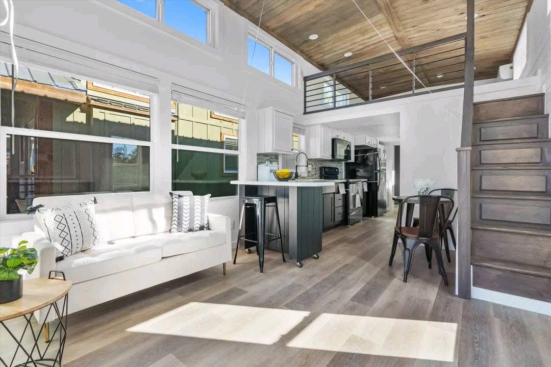 Bright, modern tiny house interior featuring a white couch with patterned cushions, a central kitchen island, wood floors, and an upper loft area, creating an open, airy feel.