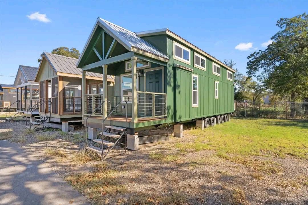 Green tiny house on wheels with a metal roof and small porch, sunny day. Surrounded by other tiny houses, trees, and open grass area.