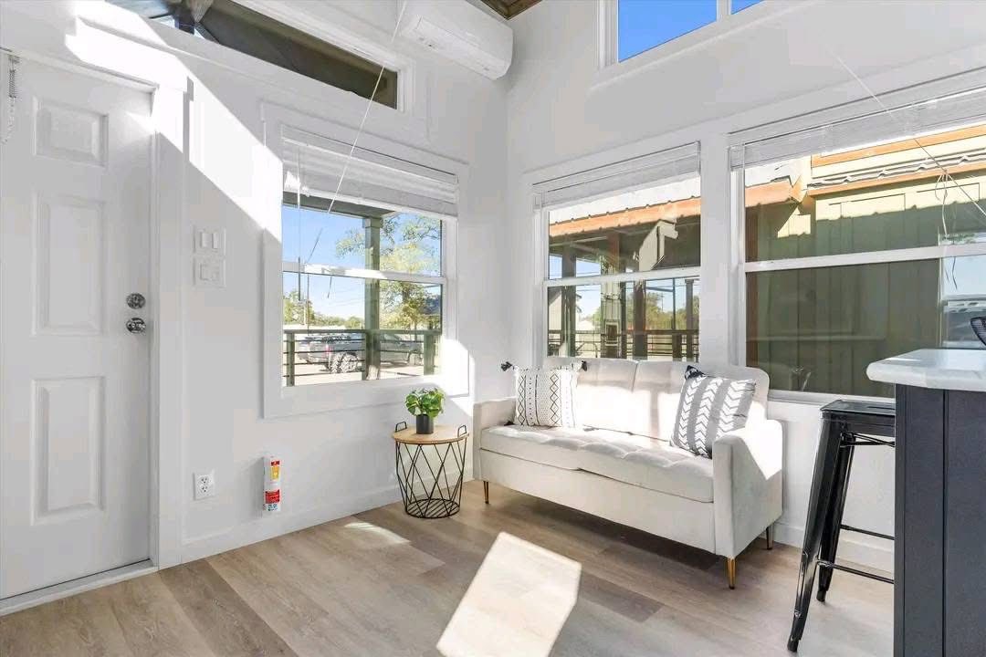 Bright, modern living space with a white couch, patterned cushions, and a round side table with a plant. Sunlight streams in through large windows.