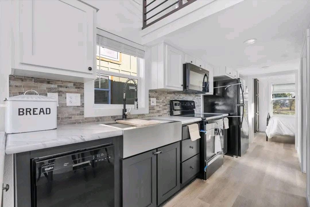 Modern kitchen with a farmhouse sink, white cabinets, and stainless appliances. A large bread box sits on the counter. Light wood floors add warmth.