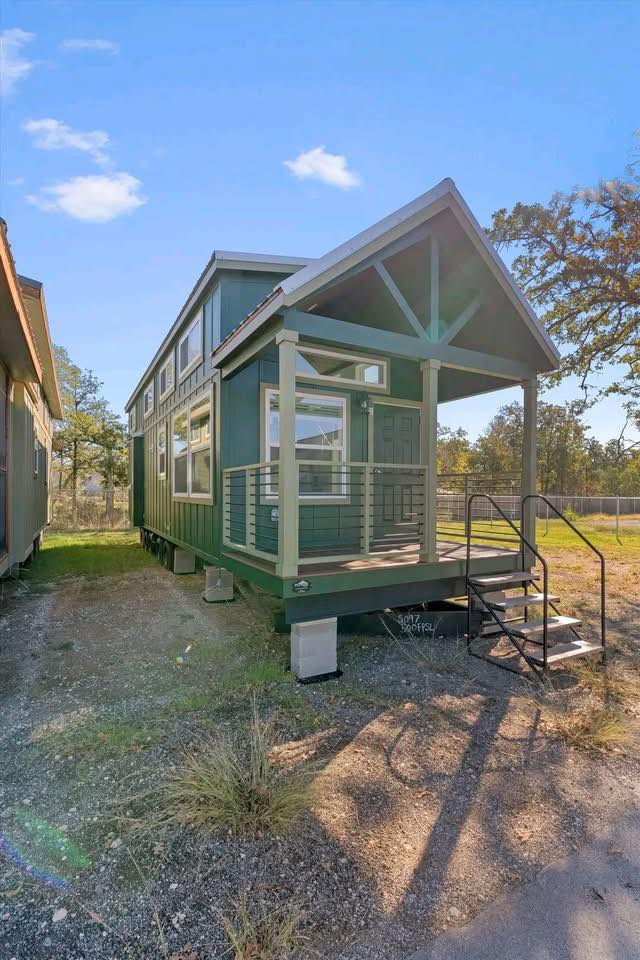 A green tiny house with a gable roof and large windows. It features a small front porch with metal railing and steps. The setting is sunny and surrounded by trees.