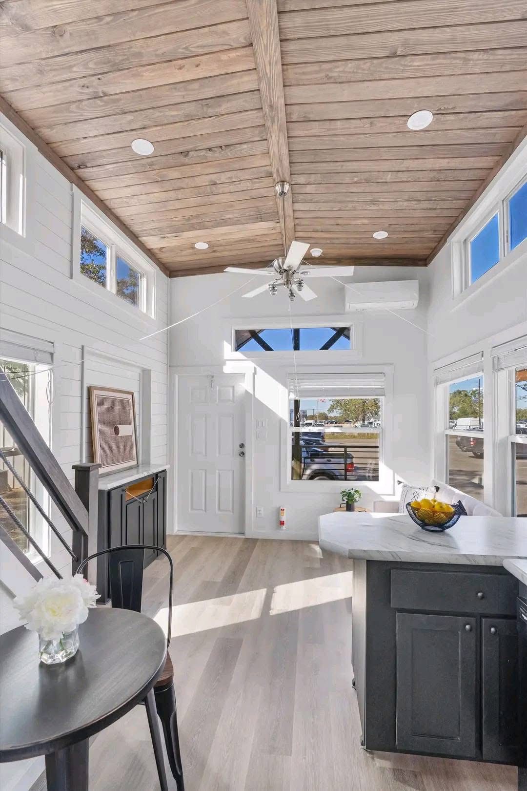 Bright and airy tiny house interior with wooden ceiling and white walls. Sunlight streams through large windows, highlighting a compact kitchen and dining area.