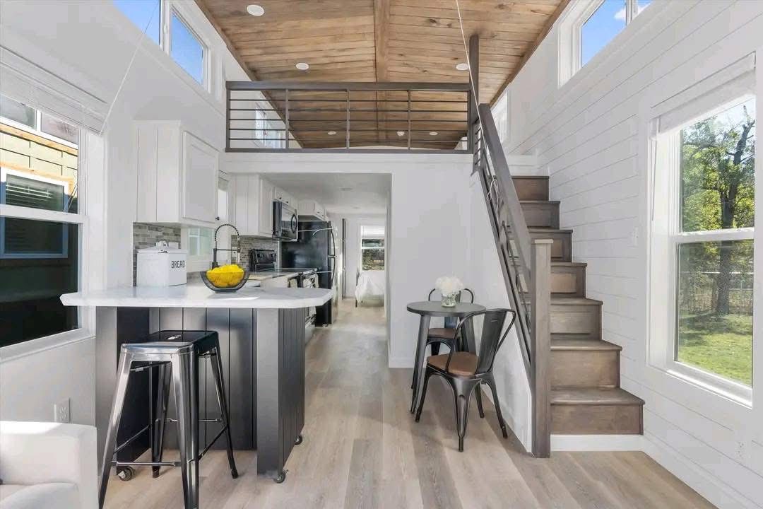 Modern tiny house interior with a cozy atmosphere. Features include a loft with wooden ceiling, sleek kitchen, bar stools at the island, and a small dining area by the stairs. Bright natural light filters through the large windows.