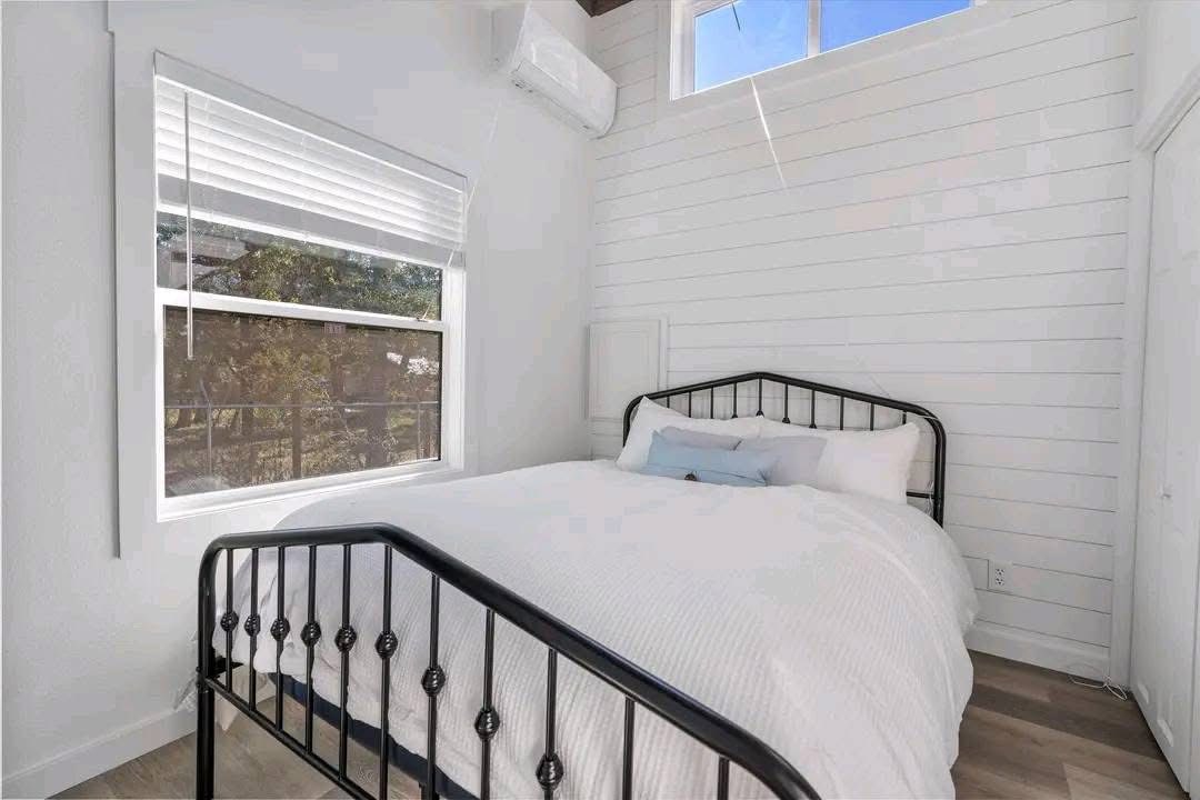 A cozy bedroom with a white shiplap wall, featuring a neatly made bed with white bedding and a black metal frame. Sunlight streams through a window with blinds, casting soft shadows. The room has a light, airy feel.