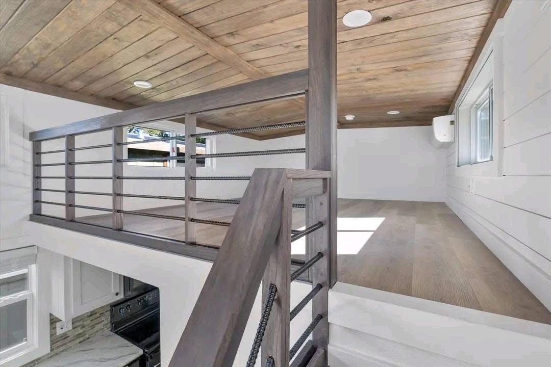 Modern loft interior with wooden ceiling and railing, light wood flooring, and white walls. Bright natural light from a window adds warmth and openness.