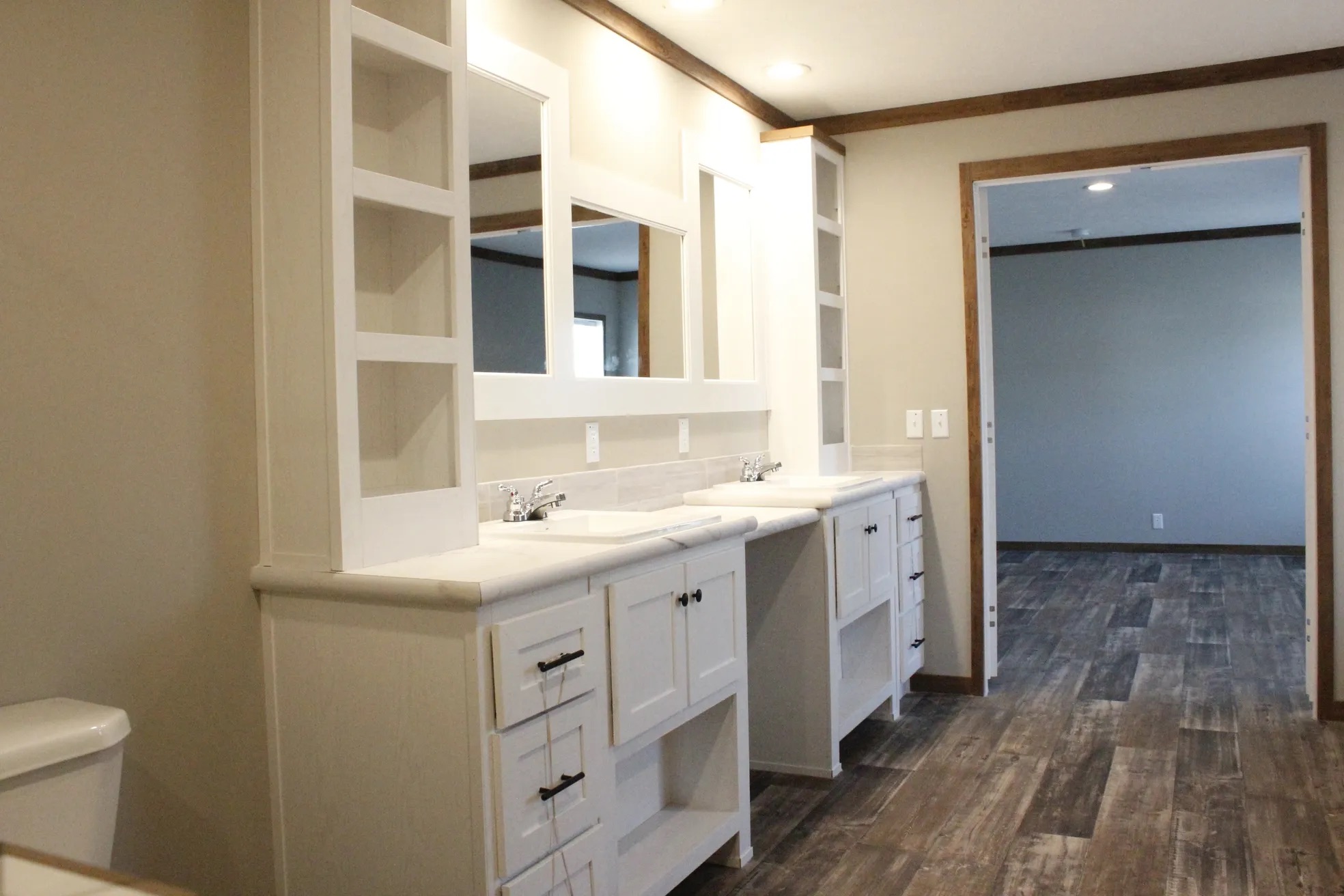 Spacious bathroom with two white vanities, rectangular mirrors, and shelving. Gray tiles cover the floor, leading into an empty adjacent room.