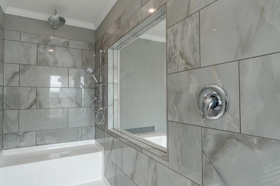 Modern bathroom with large gray marble tiles, chrome fixtures, and a rain showerhead. A built-in tub is to the left under soft, natural light.