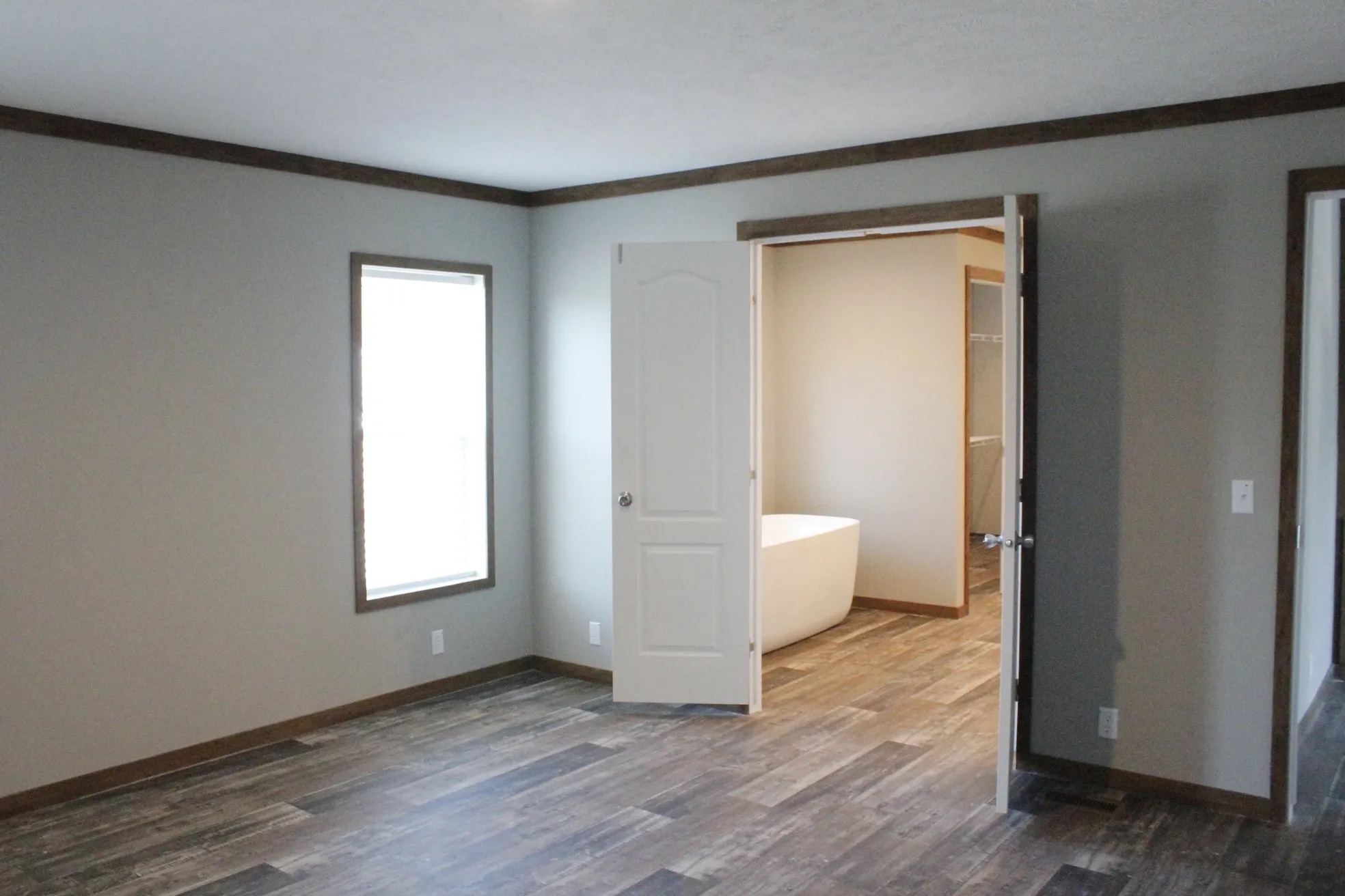 Empty room with wood flooring and light gray walls. A doorway leads to a bathroom with a white bathtub. A window provides natural light.