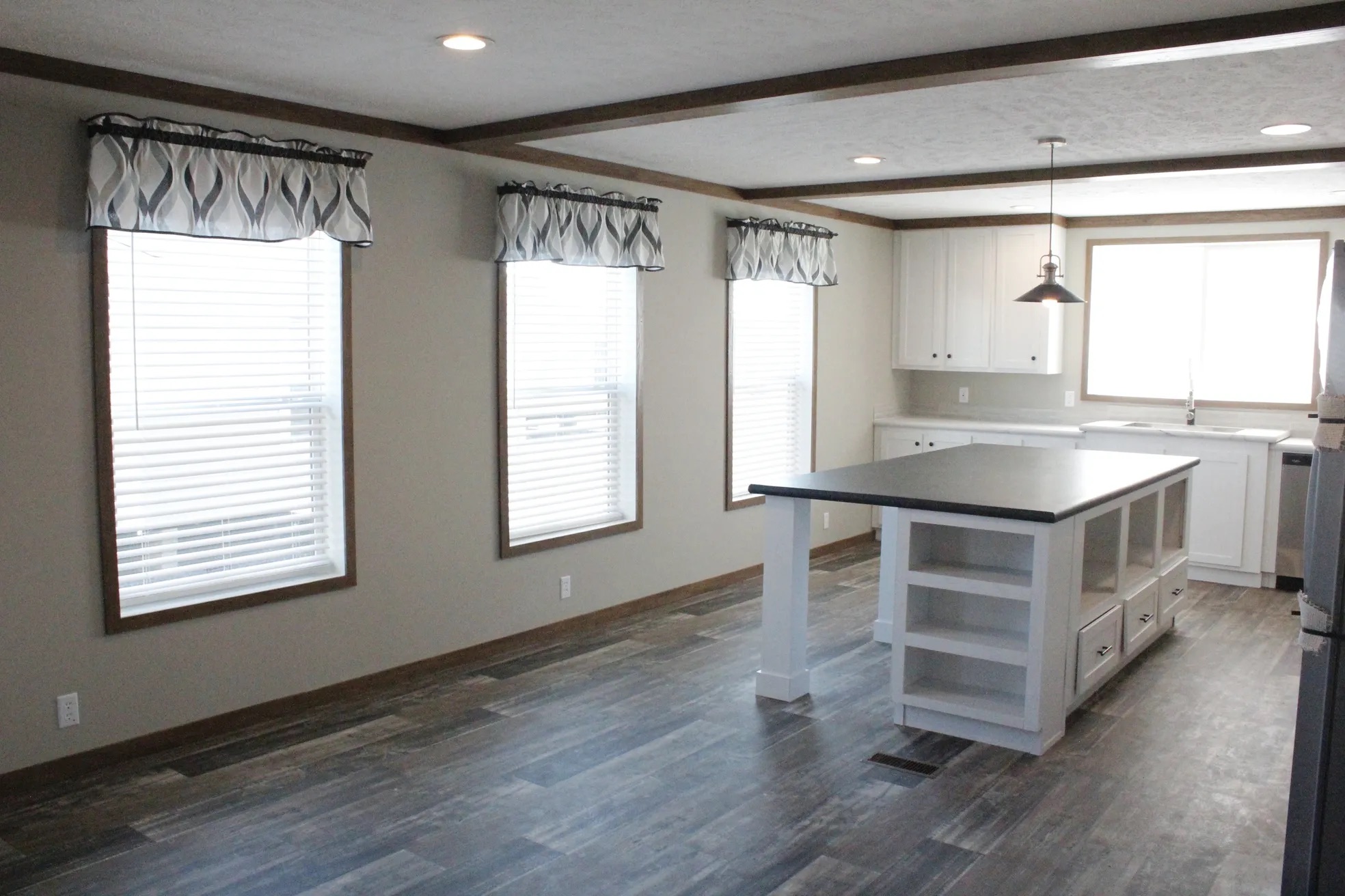 Spacious kitchen with dark wood floors, large island, and white cabinets. Three tall windows with patterned valances allow natural light to fill the room.