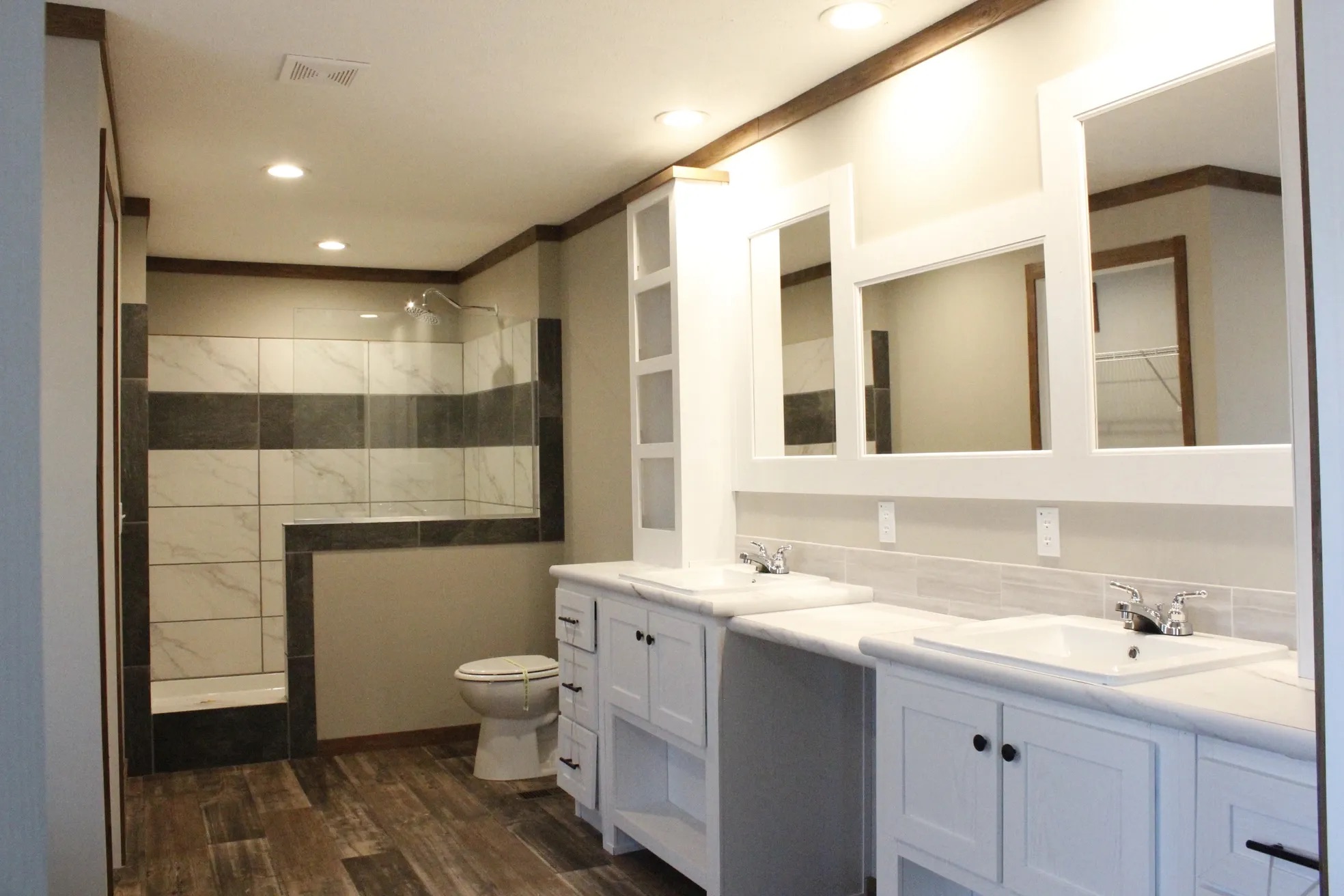 Spacious modern bathroom with dual sinks, large mirrors, and white cabinetry. A walk-in shower with gray and white tiles is in the background.