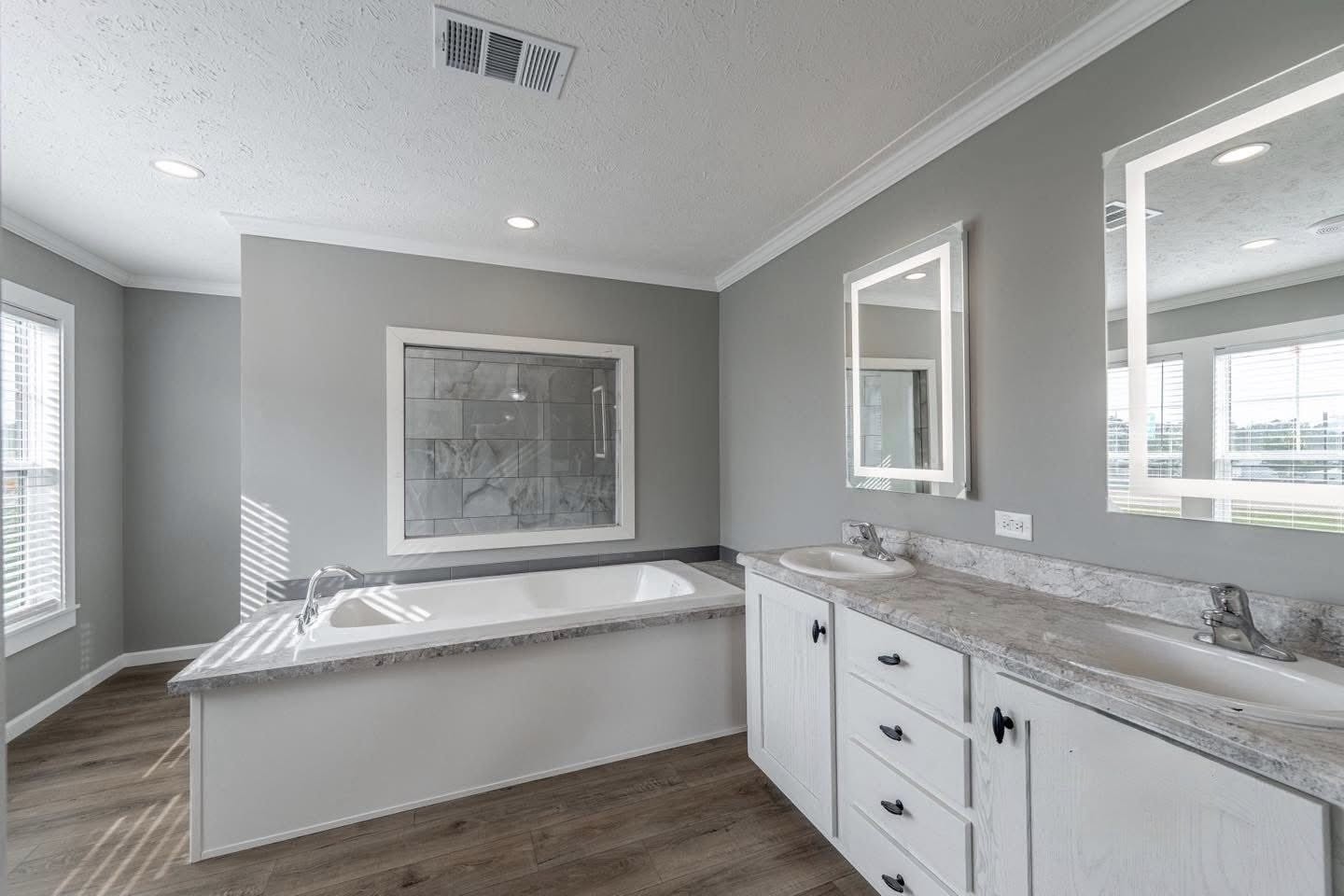Modern bathroom with a spacious bathtub, dual sinks on a white vanity, backlit mirrors, gray walls, and large windows, creating a clean, airy feel.