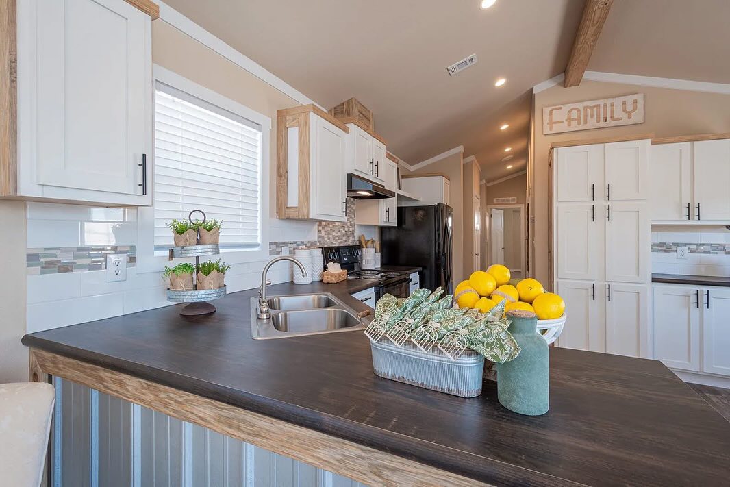 A modern kitchen features white cabinets, black countertops, and a farmhouse sink. Lemons and plants add a fresh touch. A "Family" sign hangs above.
