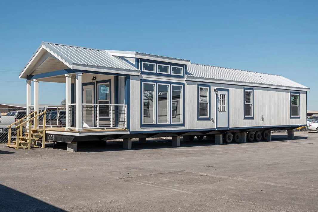 A modern mobile home with a white exterior and large windows sits on multiple wheels in a sunny parking lot, featuring a small porch with steps.