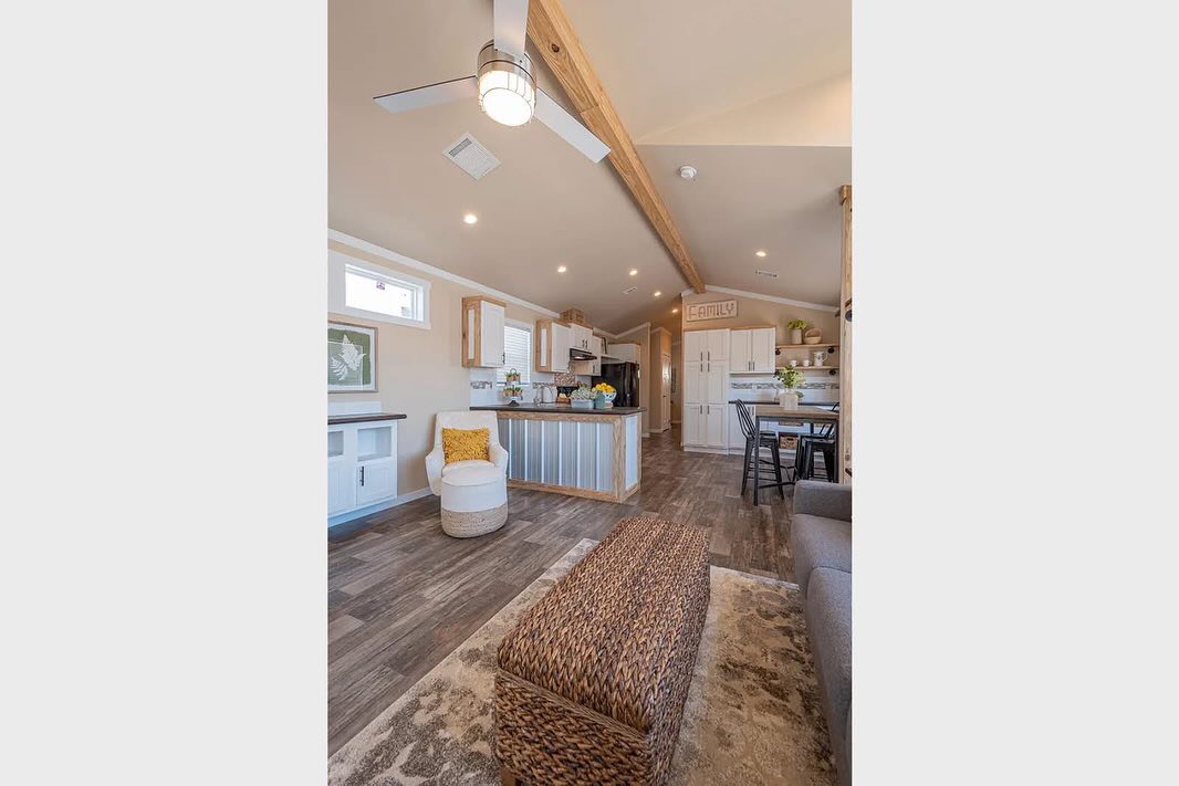 Cozy tiny house interior with a wooden ceiling beam and a ceiling fan. Features include a kitchen with modern appliances, wicker ottoman, and stylish decor.