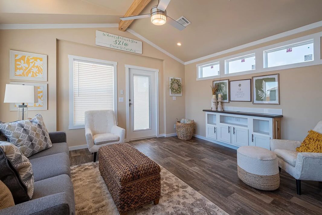 Cozy living room with beige walls, wooden floor, and vaulted ceiling. Features a gray sofa, woven ottoman, white armchairs, and decorative art. Natural light fills the space.