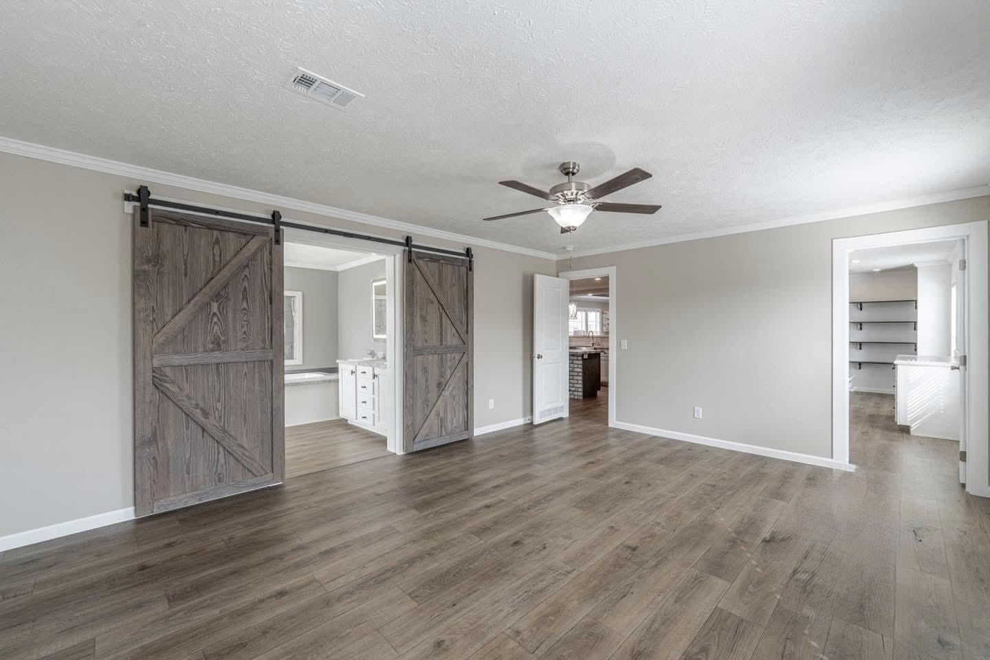 Spacious room with light wood flooring and taupe walls. Features a ceiling fan, barn doors leading to a bathroom, and an open door to a closet.