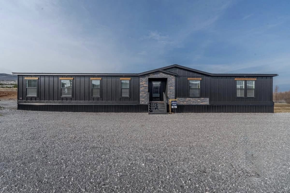 A modern, dark-paneled modular home with stone accents and multiple windows stands on a gravel lot under a clear blue sky, conveying a sleek and minimalist vibe.