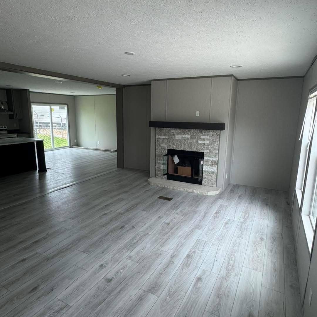 Spacious modern living room with light wood flooring, gray walls, and a stone fireplace. Natural light spills in from large windows, creating an airy feel.