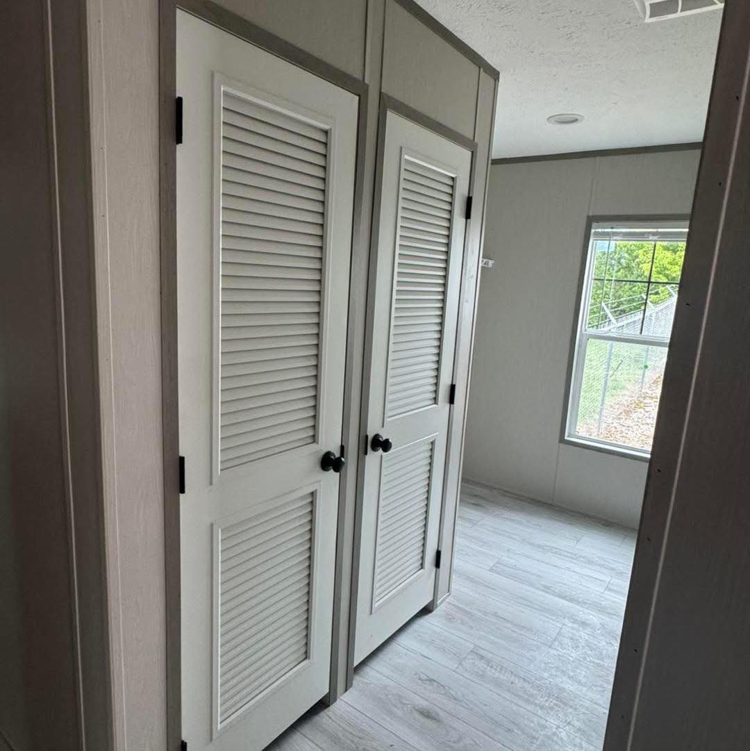 A hallway with two white louvered closet doors on the left, gray trim, and a window with a view of greenery on the right. Bright, minimalistic mood.