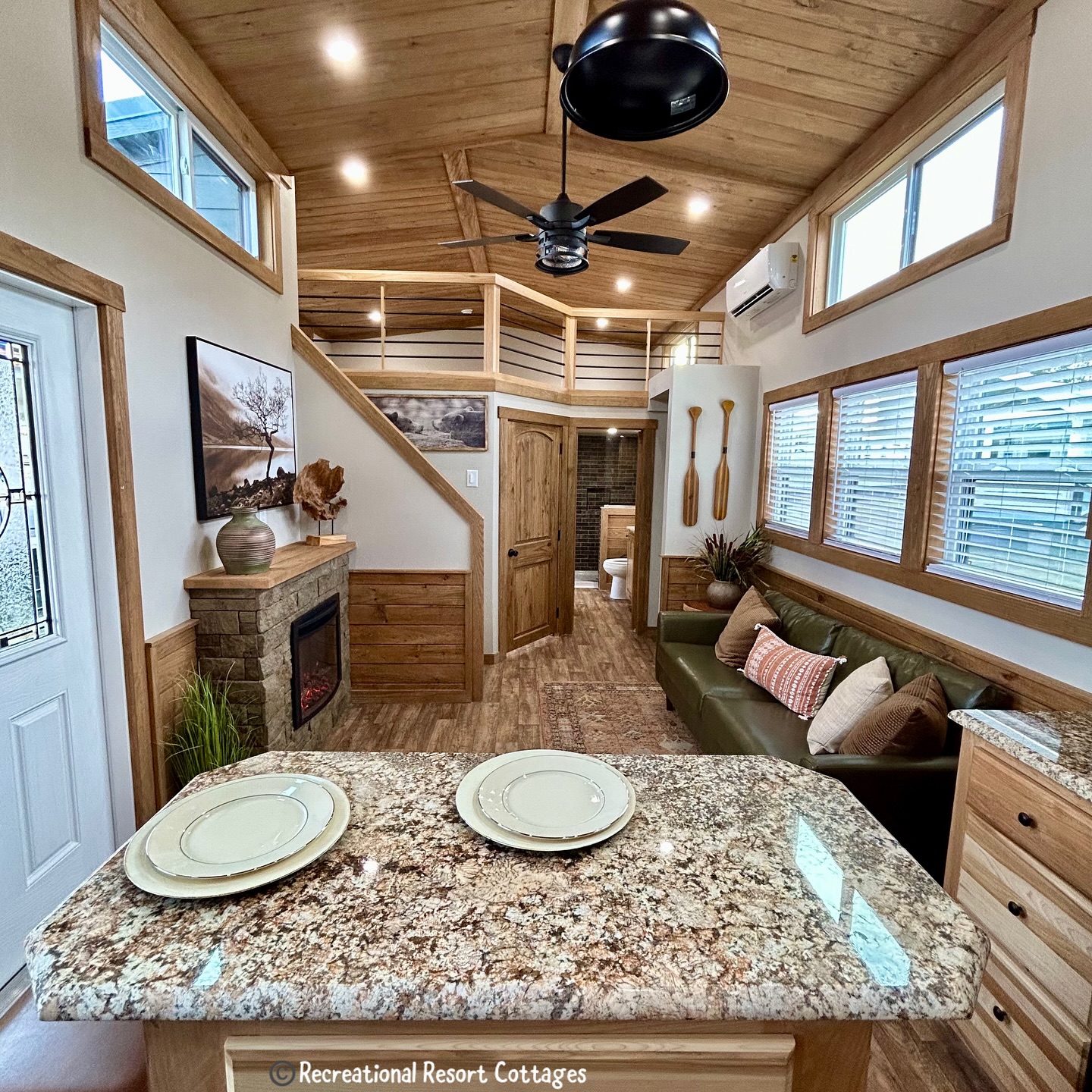 Stylish tiny home interior with wood accents. Features a granite kitchen island, cozy green sofa, stone fireplace, and ceiling fan. Warm, inviting atmosphere.