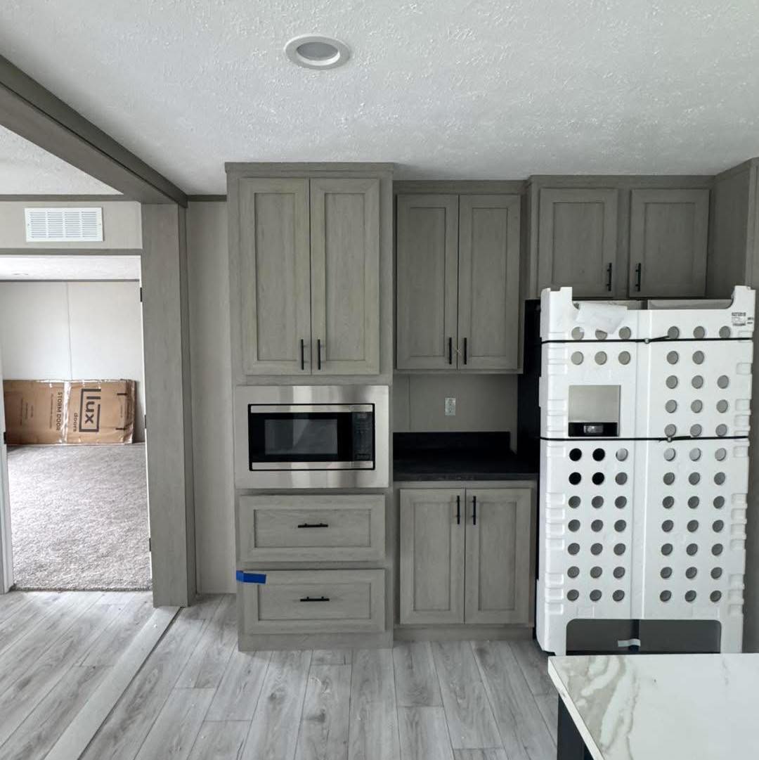 Modern kitchen scene with light wood cabinets, a stainless steel microwave, and a boxed refrigerator. Adjacent room visible with carpet flooring.