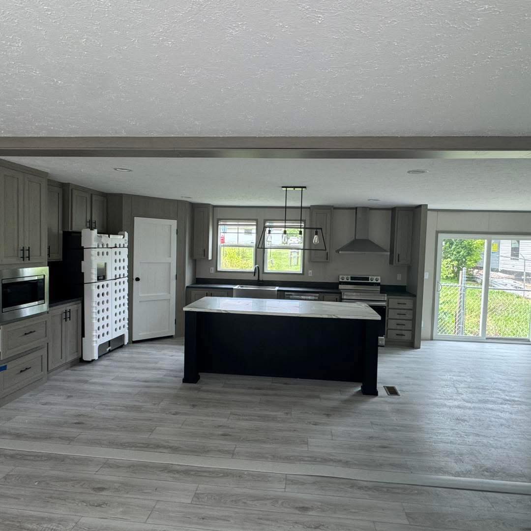 Modern kitchen with gray cabinets, marble island, stainless steel appliances, and light wood flooring. Large windows provide natural light.