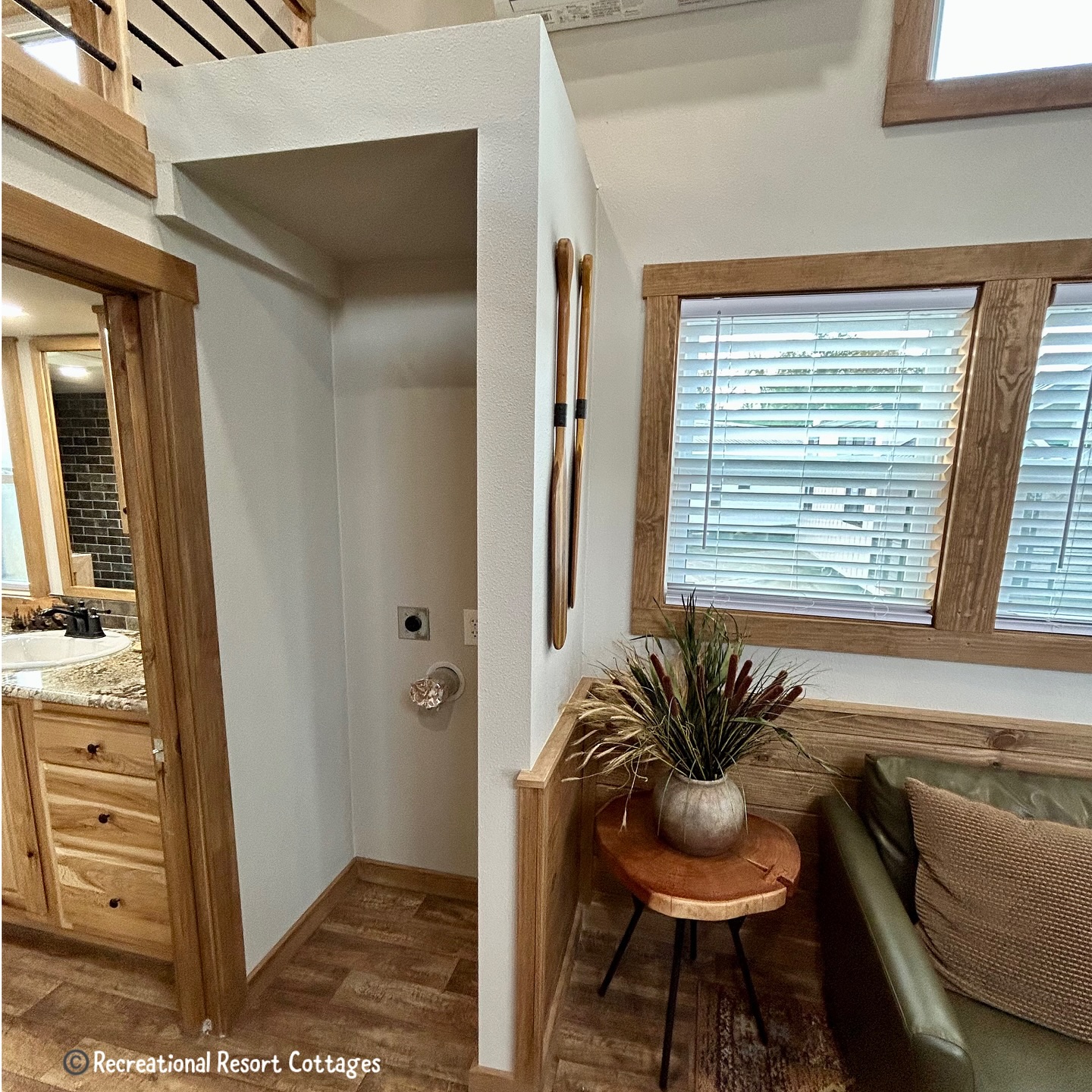 Cozy interior with wood accents. Nook for washer/dryer hookup, decorative oars on wall. Nearby, a small table holds a plant, next to a couch and large windows.