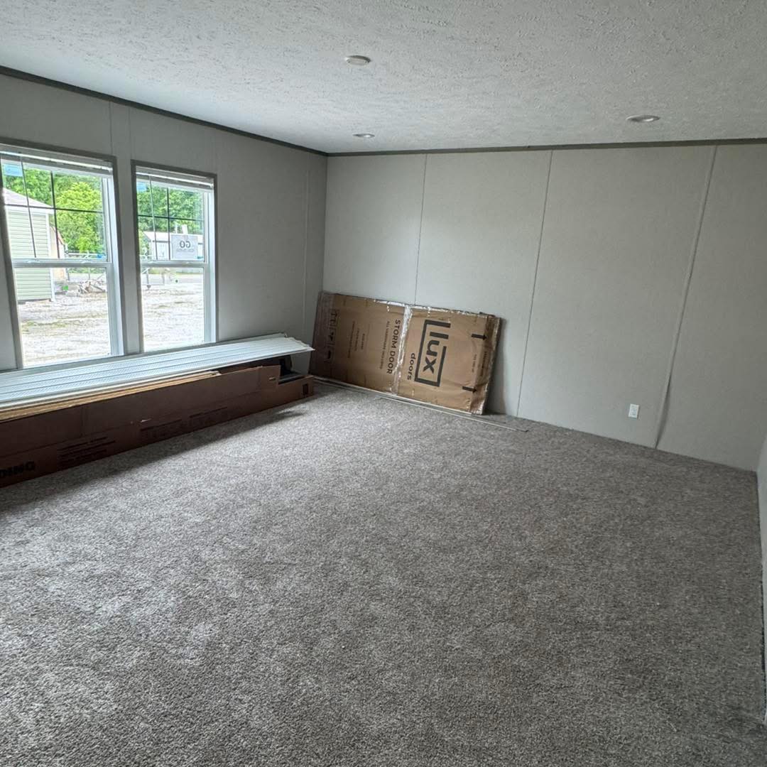 Empty room with light gray walls and beige carpet. Two large windows on the left let in natural light. Boxes and panels are stacked by the windows.