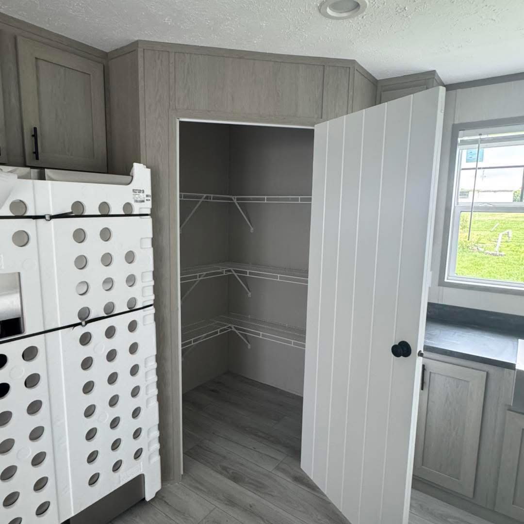 A modern kitchen corner with a light gray cabinet and an open pantry door revealing empty wire shelves. Nearby is stacked packing material. The mood is tidy and minimalistic.
