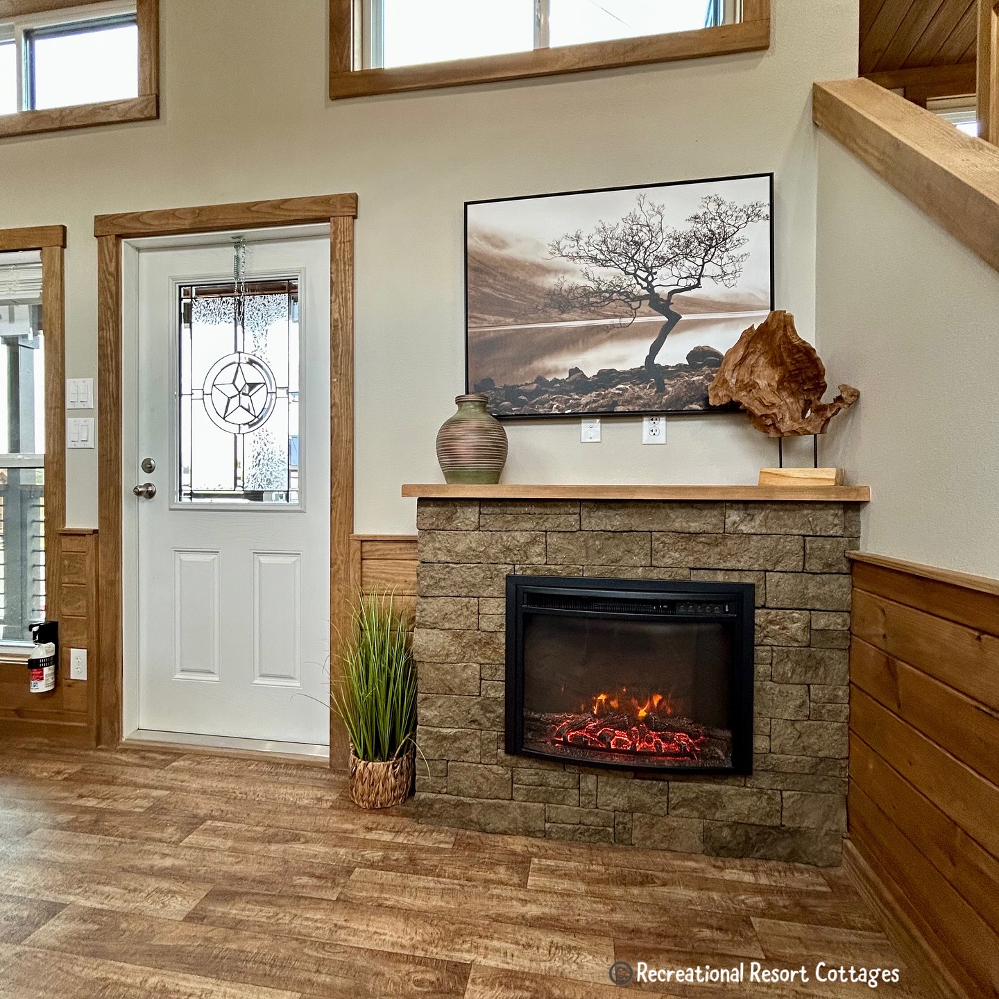 Cozy living room corner with a stone fireplace displaying an electric fire. Above, a serene tree landscape photo and rustic decor create a warm ambiance.