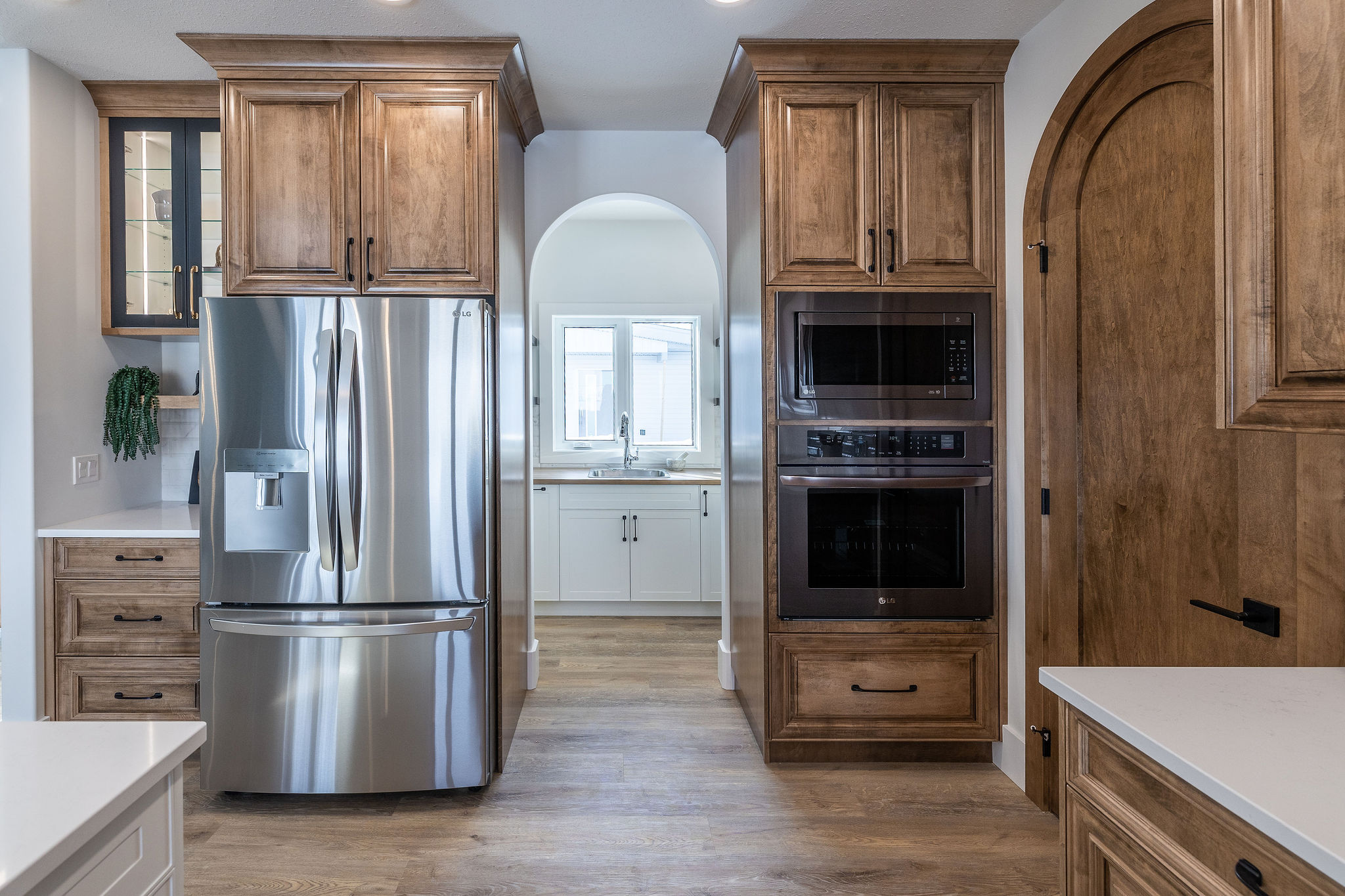 Modern kitchen with stainless steel fridge, wooden cabinets, built-in ovens, and an arched doorway leading to a bright utility space with a window.