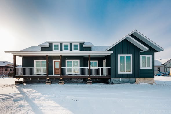 A modern prefab home on a snowy landscape, with a dark blue exterior, large windows, and a porch. The bright sky suggests a cold, clear day.