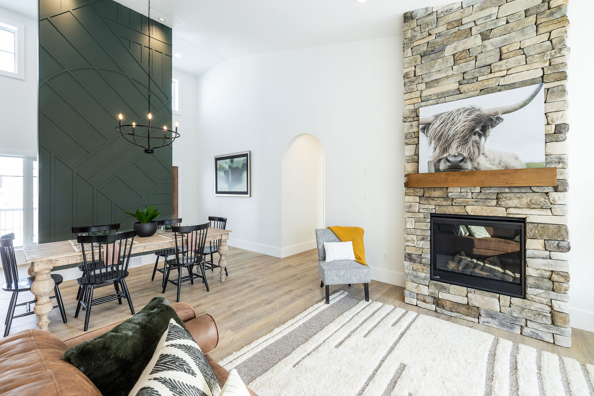 Modern living room featuring a stone fireplace with a cow photo. Wooden dining table, dark green accent wall, and cozy seating evoke a chic, rustic vibe.