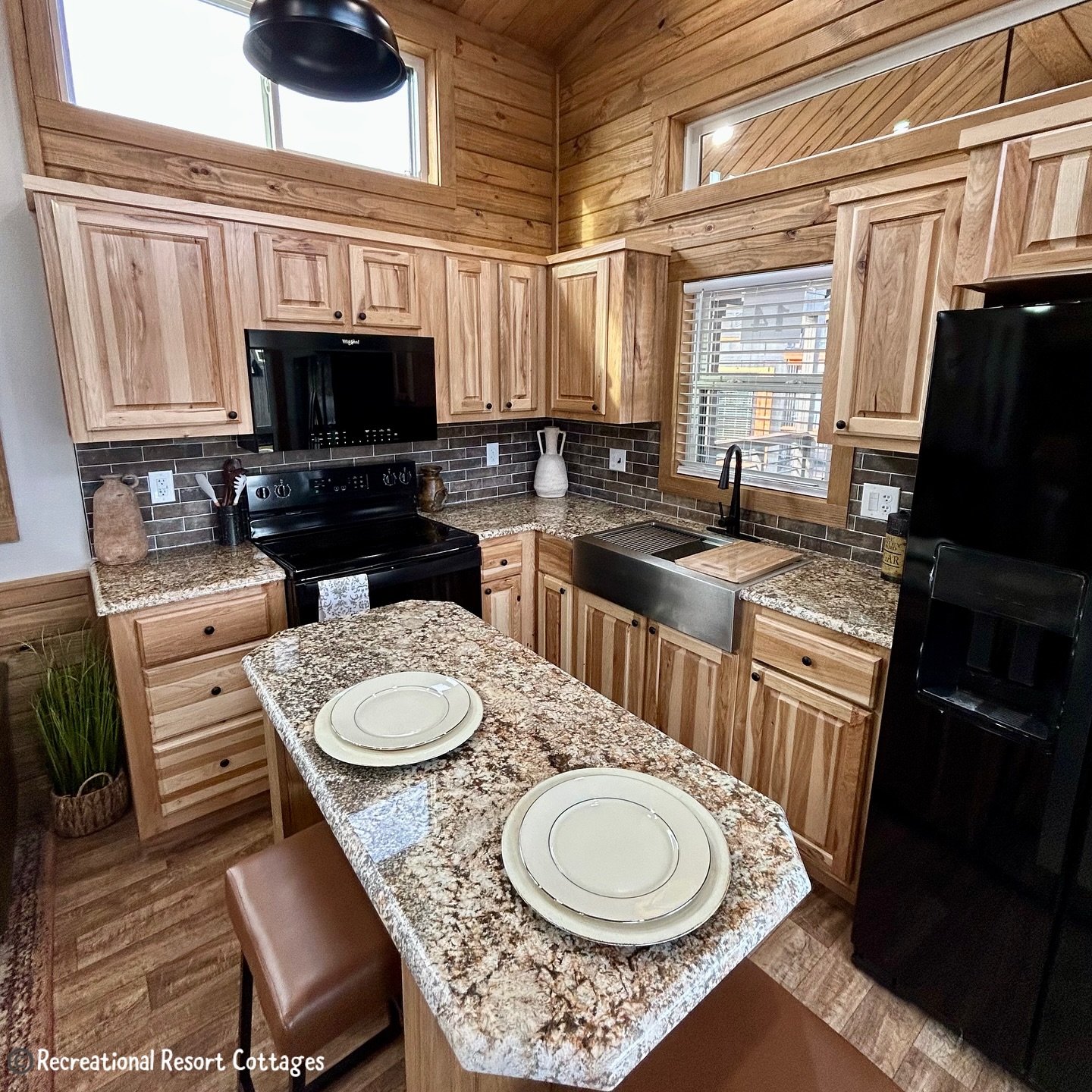 Stylish kitchen with wood cabinets, granite countertops, farm sink, and black appliances. A cozy island has two place settings, under warm lighting.