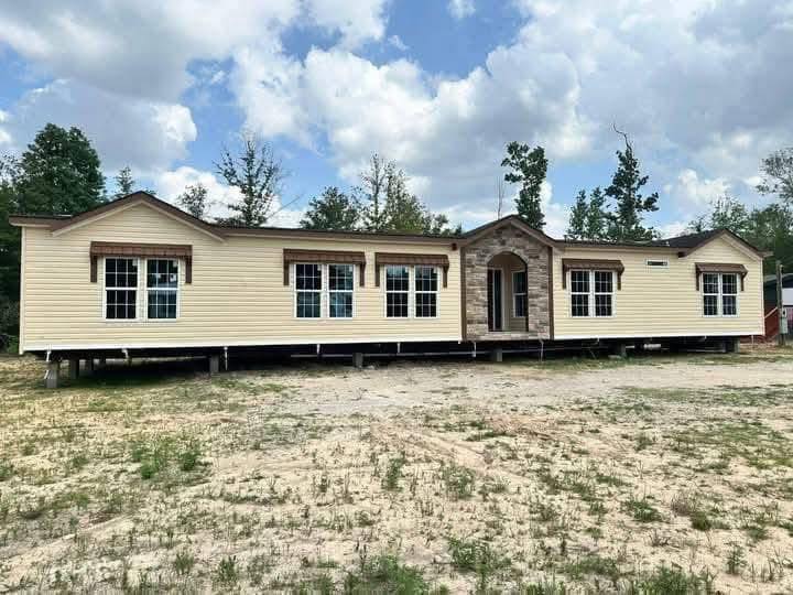 A beige, double-wide mobile home sits on a sandy lot, surrounded by trees under a partly cloudy sky. The home features multiple large windows.