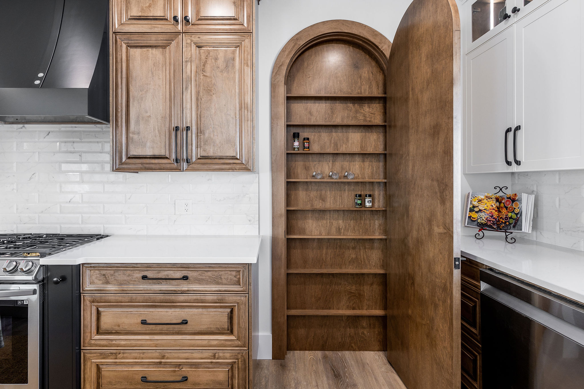 Stylish kitchen with hidden wooden pantry door ajar, white countertops, textured white tile backsplash, and sleek cabinetry. Cozy and modern atmosphere.