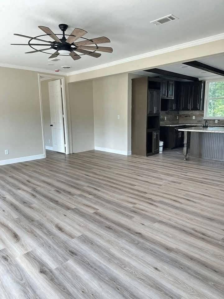 Spacious, empty room with light wood flooring, beige walls, and a ceiling fan. Open-concept layout connects to a modern kitchen with dark cabinets.