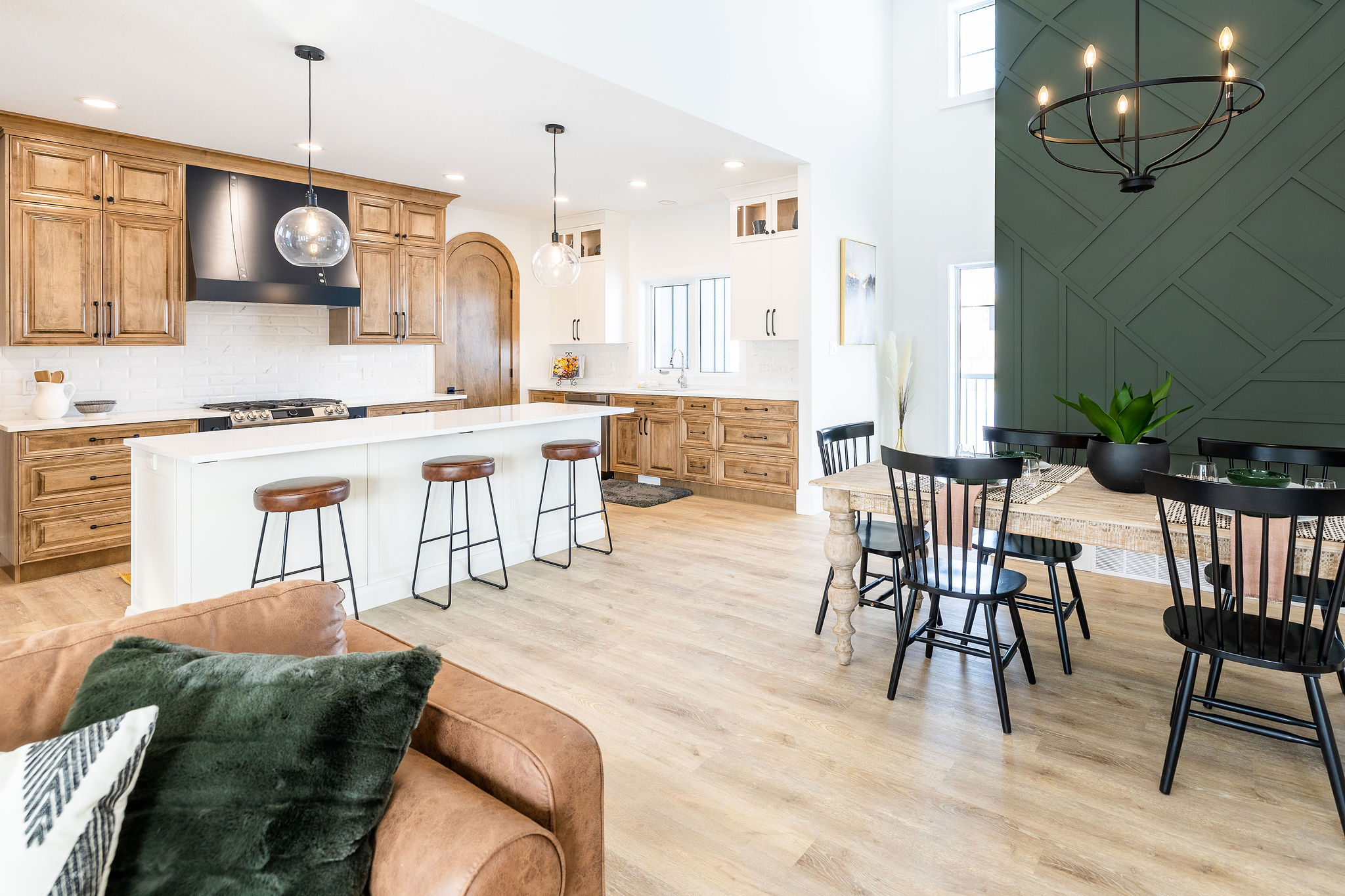 Modern kitchen and dining area with light wood floors. Features brown cabinets, a large white island with stools, and a dark green accent wall.