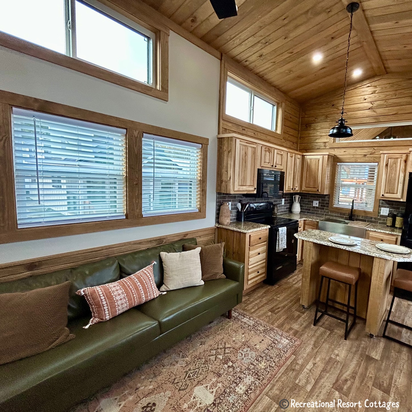 Cozy cabin interior with wooden walls and flooring. A green couch with decorative pillows faces a kitchen area with granite counters and bar stools. Warm, inviting atmosphere.