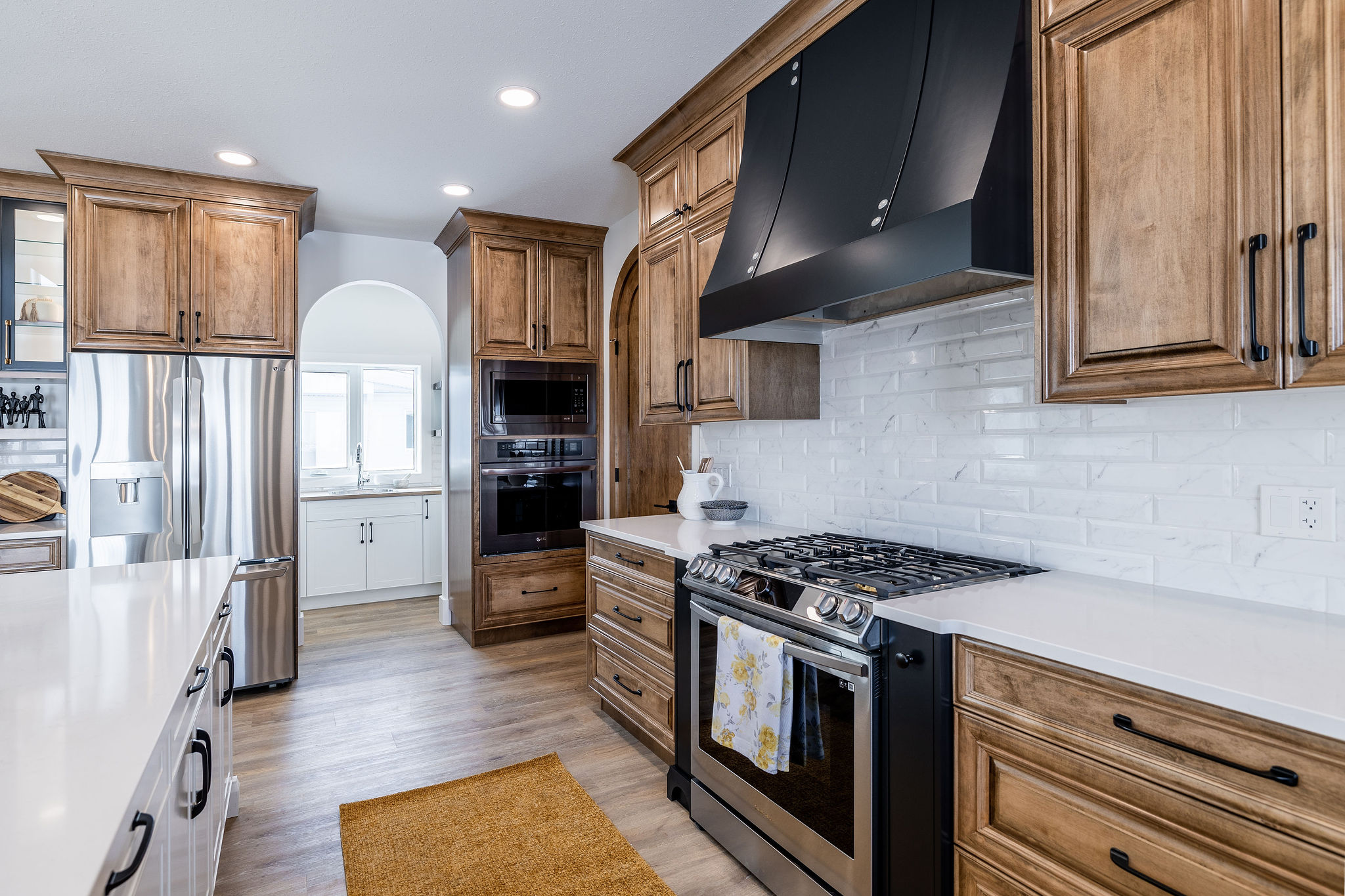 Spacious kitchen with wooden cabinets, a stainless steel fridge, white countertops, a large black range hood, and a cozy light atmosphere.