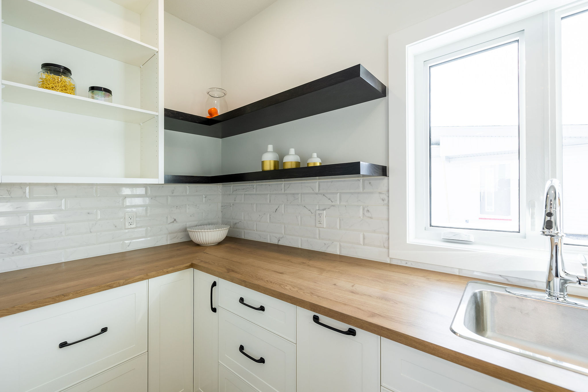 Modern kitchen corner with white cabinets, black handles, and wood countertop. Open shelves display bottles and jars. Bright window and sleek sink.
