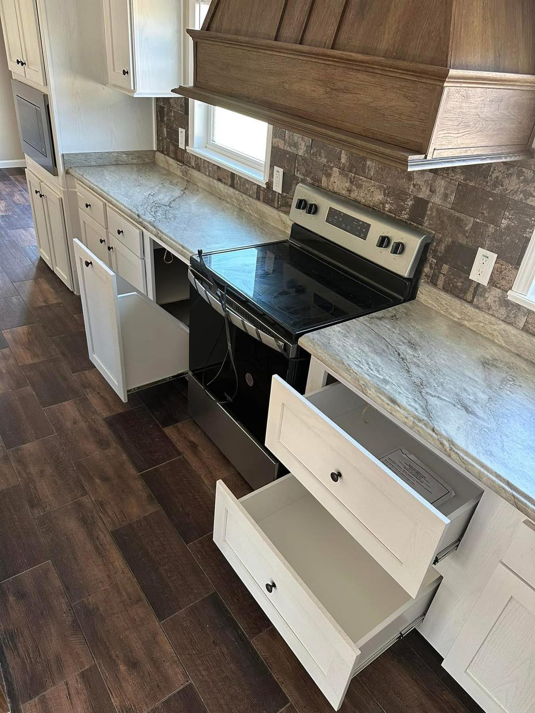 Modern kitchen with dark wood floors, open white cabinets, and drawers. Marble countertops and stainless steel oven convey a sleek, clean atmosphere.