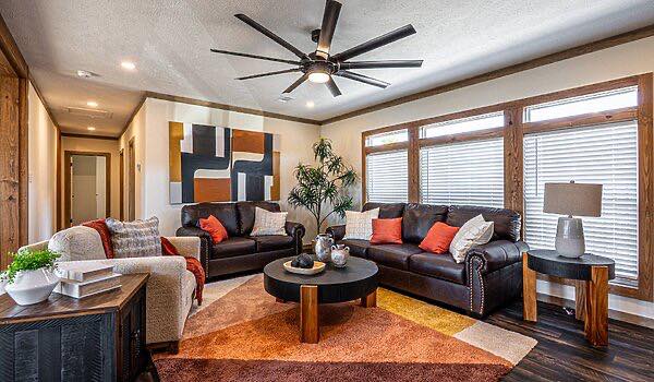 A modern living room with brown leather sofas, colorful cushions, abstract wall art, and a large ceiling fan. Bright natural light fills the space.