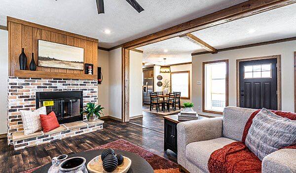 Cozy living room with a brick fireplace, wooden accents, and modern decor. A gray sofa with red pillows and a view into a dining area create a warm ambiance.