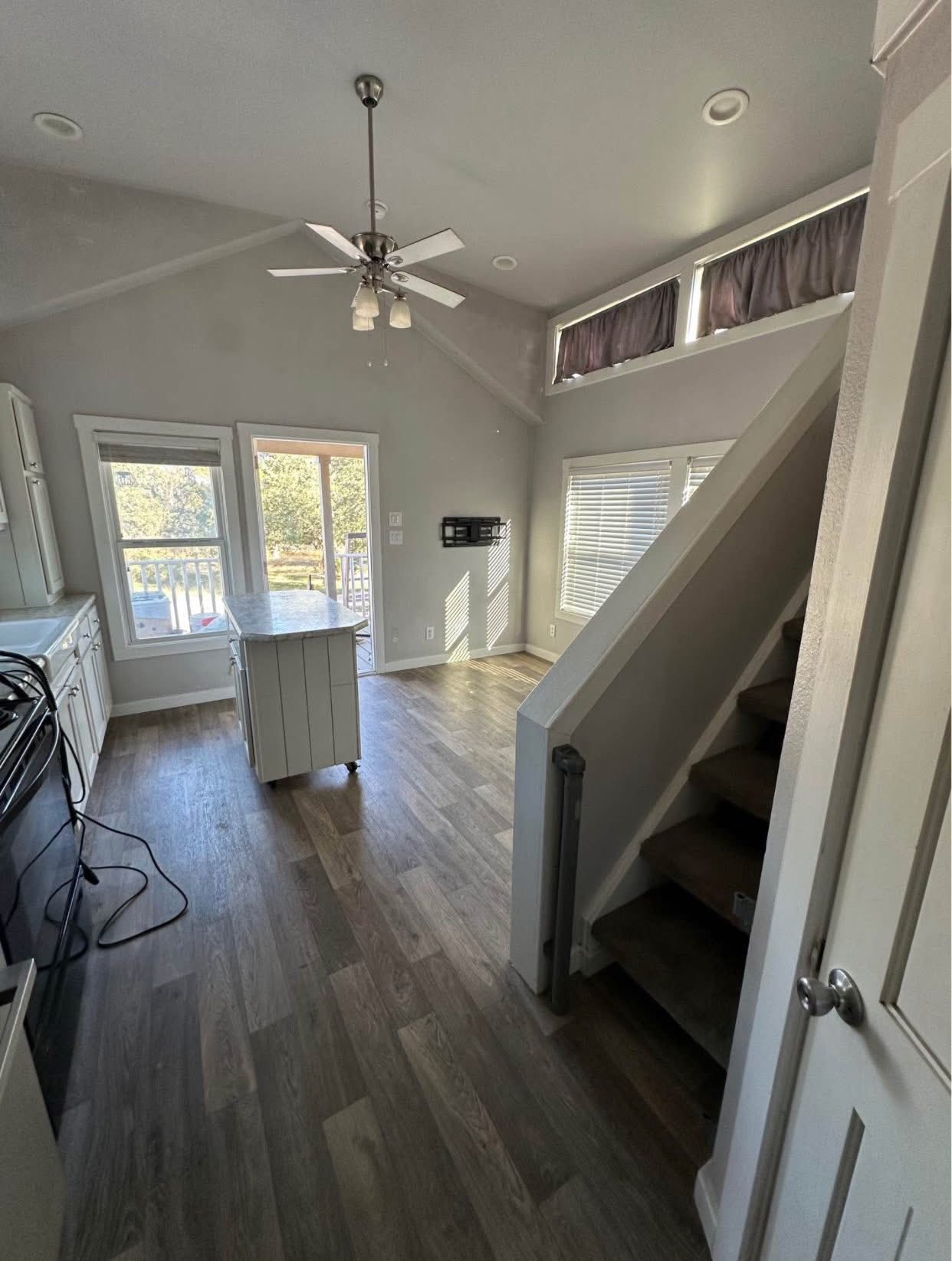 Bright, cozy room with wooden floors, large windows, and a staircase. A ceiling fan hangs above a small island, capturing a serene, airy atmosphere.