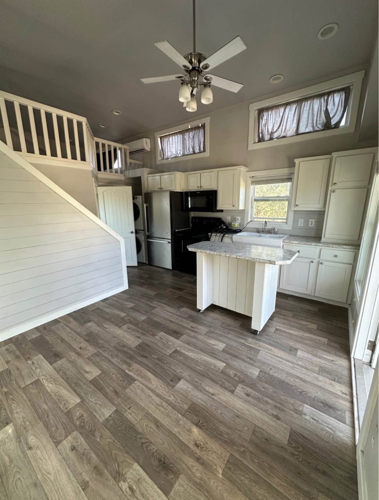A cozy kitchen with wood flooring, white cabinets, and a central island under a ceiling fan. A staircase leads to a loft with minimalist decor.